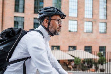 Kasklı ve sırt çantalı bir adam işe bisikletle gidiyor.