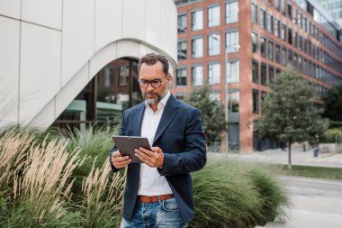 Modern ofis binasının önünde elinde tablet olan ve bir şeyler okuyan bir iş adamı..