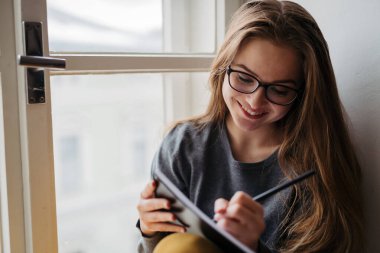 Ders kitabı okuyan kız öğrenci pencere eşiğinde oturup ders çalışıyor..