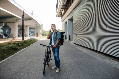 Bisikletli bir erkek, telefonla konuşuyor, bisikletinin yanında duruyor..