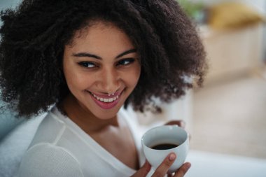 Kapalı alanda kahve içen, kameraya bakan, gülen genç bir kadının yakın plan portresi..