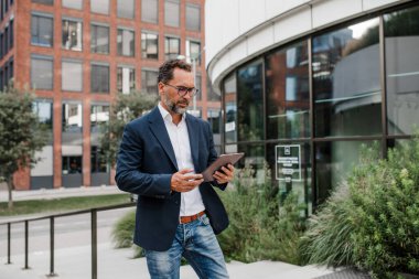 Modern ofis binasının önünde elinde tablet olan ve bir şeyler okuyan bir iş adamı..