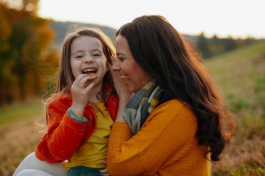 Gülen bir anne ve küçük bir kızı sonbahar doğasında bir yerde oturuyor..