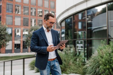 Modern ofis binasının önünde elinde tablet olan ve bir şeyler okuyan bir iş adamı..