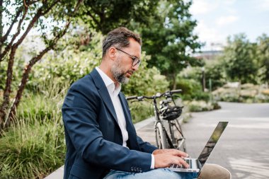 Şehir parkında çalışan erkek yönetici, telefonla konuşuyor ve dizüstü bilgisayarda daktilo yazıyor..
