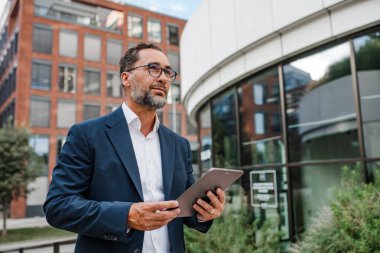 Modern ofis binasının önünde elinde tablet olan ve bir şeyler okuyan bir iş adamı..