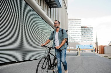 Sokakta bisiklet süren, sırt çantası takan bir banliyö. Kameraya bakıp gülümsüyor..