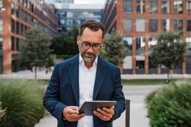 Modern ofis binasının önünde elinde tablet olan ve bir şeyler okuyan bir iş adamı..