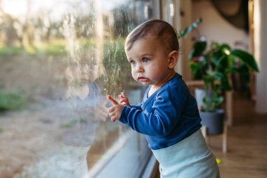 Küçük bir çocuk, üzerinde parmak izleri olan pencereden dışarı bakıyor..