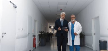 Yönetici doktorla koridorda yürüyor, tabletteki belgeyi gösteriyor. Hastane ayarları.