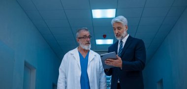 Yönetici doktorla koridorda yürüyor, tabletteki belgeyi gösteriyor. Hastane ayarları.