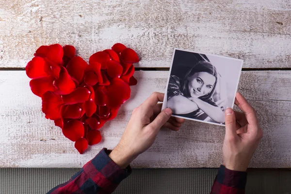 Mans hands holding his girlfriends photo. Red rose petal heart. — Stockfoto