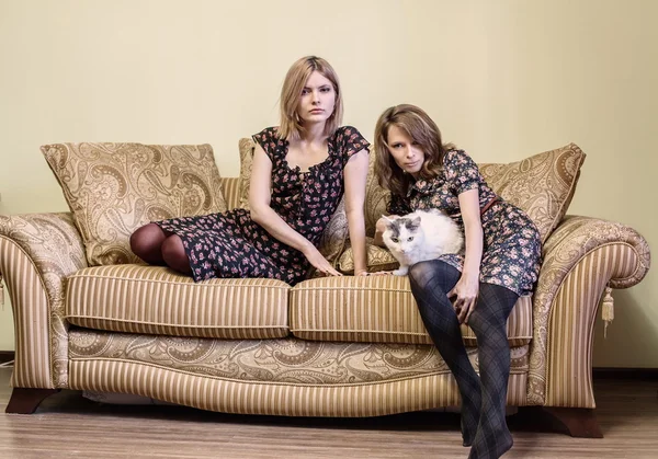 Two beautiful girls in dresses sitting on a sofa. — Stock Photo, Image