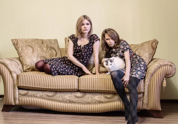 Duas meninas bonitas em vestidos sentados em um sofá . Fotos De Bancos De Imagens