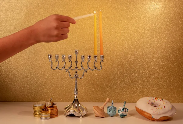 Day 2 of jewish religious holiday Hanukkah with child's hand lighting 2nd candle in traditional chandelier menorah on golden glittering background with other traditional objects around...