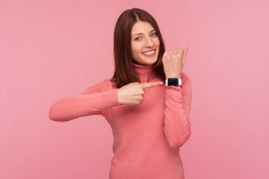 Kahverengi saçlı, pembe kazaklı mutlu bir kadın bileğini işaret ederek kameraya dişlek tebessümle bakıyor. Saate bak! Pembe arka planda kapalı stüdyo çekimleri