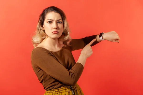 We have no time! Portrait of serious boss woman with blonde hair in brown blouse pointing her finger at the smartwatch and getting angry. Indoor studio shot isolated on red background