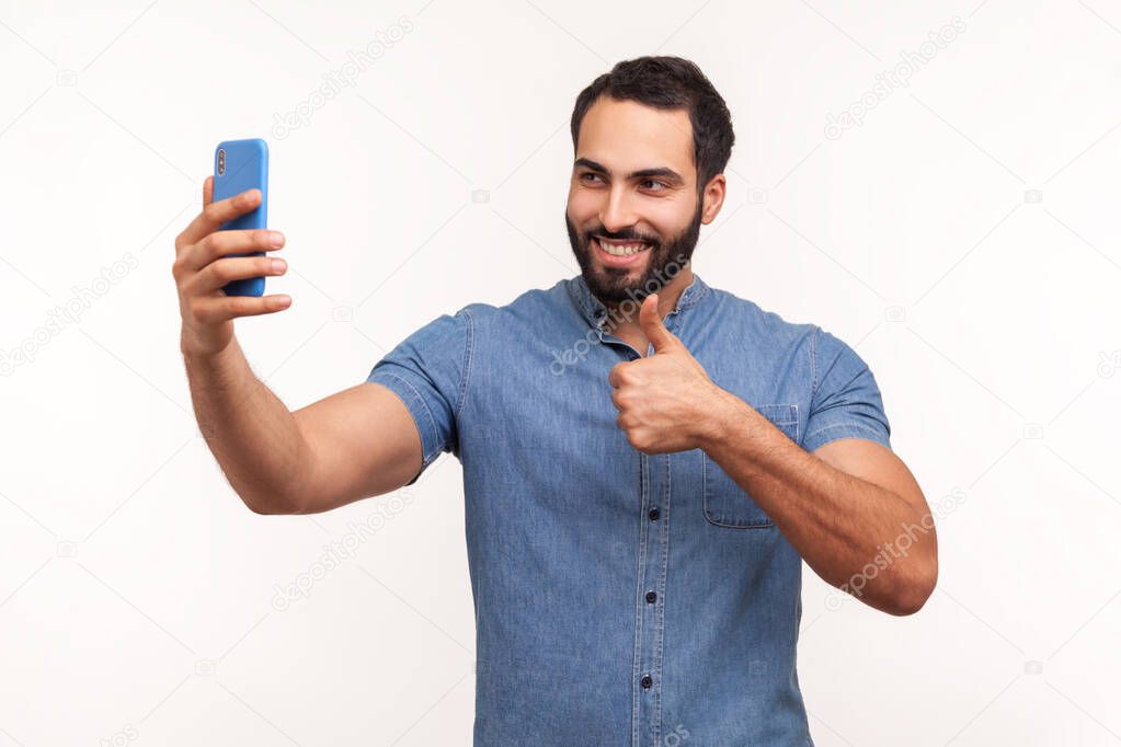 Extremely satisfied happy bearded man showing thumbs up and smiling looking at camera of his smartphone, blogging. Indoor studio shot isolated on white background