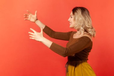 Sarışın, gülümseyen, kollarını açıp sarılan ve yeni bir arkadaşlığa hazır güzel ve açık ellerin portresi. Kırmızı arkaplanda kapalı stüdyo çekimleri