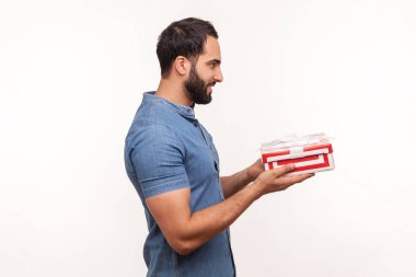 Profil portresi gülümseyen mavi gömlekli sakallı adam hediyesini çizgili hediye kutusunda tutuyor yıldönümünü kutluyor, tatillere hazırlanıyor. Beyaz arka planda izole edilmiş kapalı stüdyo görüntüleri