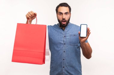 Elinde kağıt torbalar ve boş akıllı telefonlar tutan şok olmuş bir adam kolay online alışveriş ve büyük indirimler ve reklamlarla şaşırmış. Beyaz arka planda izole edilmiş kapalı stüdyo görüntüleri