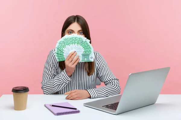 刺激的なブルネットの女性が縞模様のシャツを着てオフィスでノートパソコンに座って 数百ユーロの紙幣を見て 高い給与にショックを受けた ピンクの背景に隔離された屋内スタジオショット — ストック写真