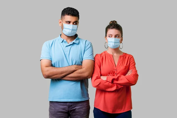 Portrait of upset couple with surgical medical mask standing together with crossed hands, looking sideways at each other with resentful glance, suspicion. isolated on gray background, indoor studio