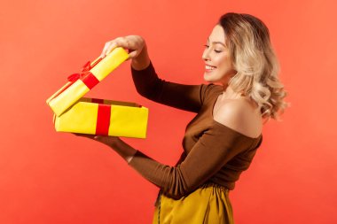 Happy beautiful woman unboxing big yellow present and looking inside with satisfied smile on face, enjoying present. Indoor studio shot isolated on orange background clipart