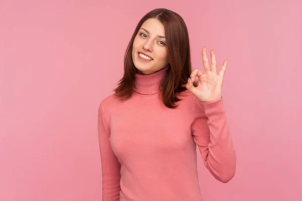 大丈夫よ ピンクのセーターの陽気なブルネットの女性は おもちゃの笑顔でカメラを見て手でOkジェスチャーを示す承認 ピンクの背景に隔離された屋内スタジオショット — ストック写真