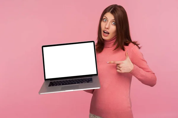 ピンクのセーターを着た幸せな驚きのブルネットの女性は 空のラップトップ画面を指して Web広告のための空白の表示を示しています ピンクの背景に隔離された屋内スタジオショット — ストック写真