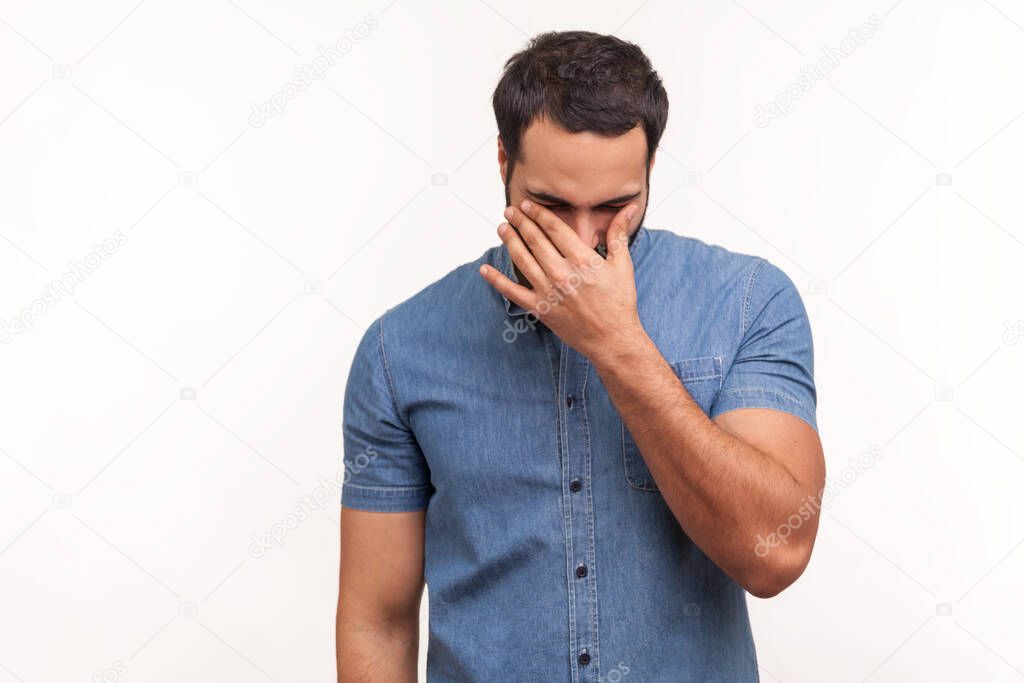 Depressed desparated man in blue shirt rubbing tired eyes, crying because of hopelessness and loneliness, nervous breakdown. Indoor studio shot isolated on white background