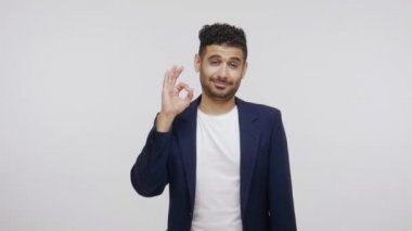Takım elbiseli, sakallı mutlu bir adam iyi bir jest yapıyor, işini onaylıyor, gurur duyuyor ve iyi sonuçlarla ve başarıyla eğleniyor. Gri arka planda izole edilmiş kapalı stüdyo görüntüleri