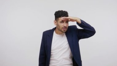 İş elbisesi giymiş kafası karışmış sakallı bir adam el ele tutuşmuş uzaktan bir şey arıyor, dikkatle izliyor. Gri arka planda izole edilmiş kapalı stüdyo görüntüleri