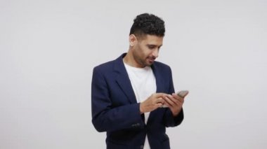 Takım elbiseli pozitif sakallı iş adamı akıllı telefondan kayıyor ve gülümsüyor, online alışveriş yapıyor ya da komik videolar izliyor. Gri arka planda izole edilmiş kapalı stüdyo görüntüleri