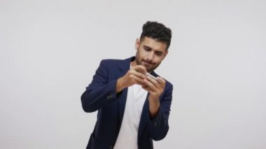 Takım elbiseli çocuksu sakallı bir adam akıllı telefondan aksiyon oyunları oynuyor, sanal gerçeklik, oyun. Gri arka planda izole edilmiş kapalı stüdyo görüntüleri