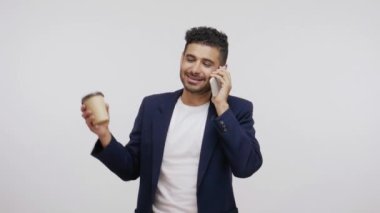Zarif takım elbiseli sakallı iş adamı cep telefonundan konuşuyor ve sıcak içecek içiyor, elinde kağıt bardak, paket kahve servisi yapıyor. Gri arka planda izole edilmiş kapalı stüdyo görüntüleri