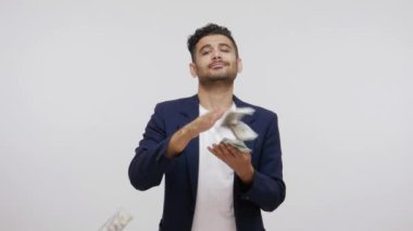 Ticari sakallı zengin adam, takım elbiseli, etrafa para saçıyor, kameraya tatmin olmuş bir ifadeyle bakıyor, yüksek maaşla övünüyor. Gri arka planda izole edilmiş kapalı stüdyo görüntüleri