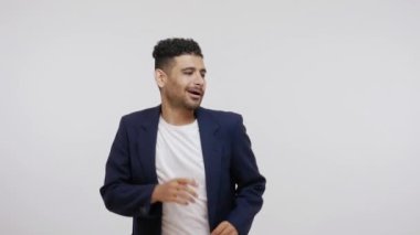 Takım elbiseli olumlu sakallı adam dans ederek ve ritme göre hareket ederek ellerini ovuşturarak dublaj yapıyor. Gri arka planda izole edilmiş kapalı stüdyo görüntüleri