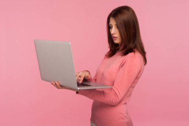 Ciddi odaklı, pembe kazaklı, kahverengi saçlı bir kadın dizüstü bilgisayarda çalışıyor, projesini bitiriyor, uzaktan kumandalı, serbest çalışıyor. Pembe arka planda kapalı stüdyo çekimleri