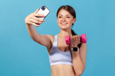 Beyaz bluzlu heyecanlı bir spor kadını dambılla selfie çekiyor, akıllı telefondan akıllı telefon uygulaması üzerinden göstergelerini izliyor, blog yazıyor. Mavi arka planda kapalı stüdyo çekimi