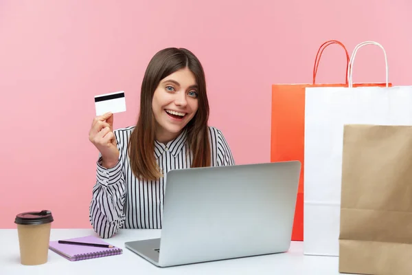 職場に座ってオンラインで買い物をするクレジットカードを示す幸せな満足女性オフィスワーカー テーブルの上に立って紙のショッピングバッグ ピンクの背景に隔離された屋内スタジオショット — ストック写真