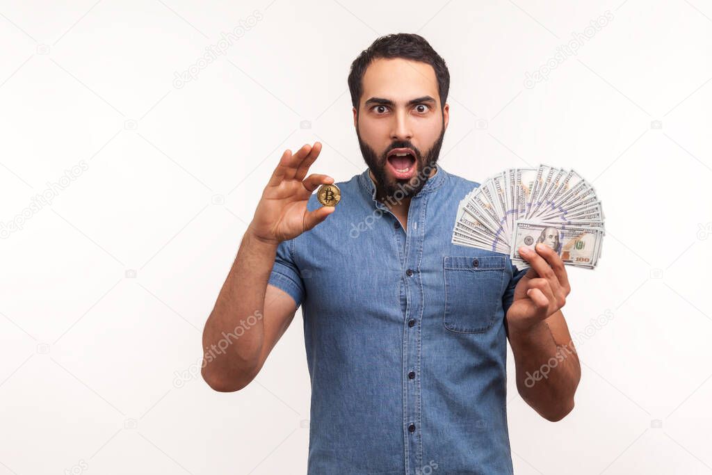 Excited bearded man holding in hands and showing dollar bills and golden coin of crypto currency, shocked by value of bitcoin. Indoor studio shot isolated on white background