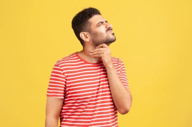 Unhappy bearded man in striped t-shirt grimacing touching his neck, feeling pain while swallowing, sore throat, risk of choking or anaphylactic shock. Indoor studio shot isolated on yellow background clipart