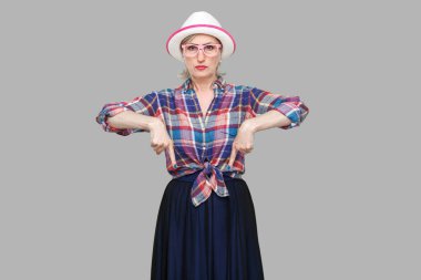 Burada ve şimdi. Ciddi modern stil olgun kadın portresi. Sıradan tarzda. Şapkası ve gözlükleriyle dikilip kameraya bakıyor. Stüdyo çekimi gri arkaplanda izole edildi.