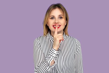 Güzel, mutlu, klasik çizgili tişörtlü bir öğretmen kırmızı dudakta parmağını tutuyor ve gülümsüyor, işaret ediyor, kapa çeneni, sınıfta konuşmayı kes ve sessiz ol. Mor arka planda izole edilmiş kapalı stüdyo çekimi