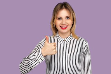 Onay işareti gibi! Parlak kırmızı dudaklı neşeli güzel bir kadın. Başparmaklarını kaldırıyor. Heyecanlı gözlerle bakıyor. Stüdyo çekimi mor arkaplanda izole edildi