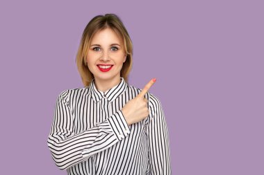 Çizgili bluzlu kırmızı dudaklı harika bir kadın boş fotokopi alanını işaret ediyor ve samimi bir şekilde gülümsüyor, reklam içeriği için yan boş duvarı gösteriyor. Mor arka planda kapalı stüdyo