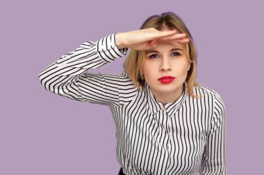 Çizgili bluzlu kırmızı dudaklı, meraklı genç bir kadın avucunu başının üstünde tutuyor ve dikkatle uzaklardan bakıyor, uzun mesafeden beklentilerle bakıyor. Mor arka planda izole edilmiş kapalı stüdyo çekimi