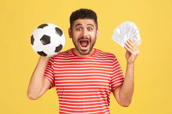 ストライプのTシャツを保持サッカーボールと100ドル札のひげを持つ非常に興奮した幸せな男は カメラを見て叫んで 賭けと勝利 黄色の背景に隔離された屋内スタジオショット — ストック写真