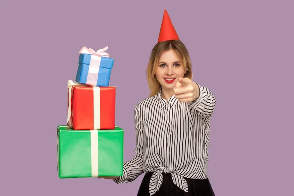 善良的快乐女人 头上挂着滑稽的派对圆锥 指着相机 拿着许多礼品盒 选择你来收到礼物和奖金 祝贺你的生日 工作室拍摄紫色背景 — 图库照片